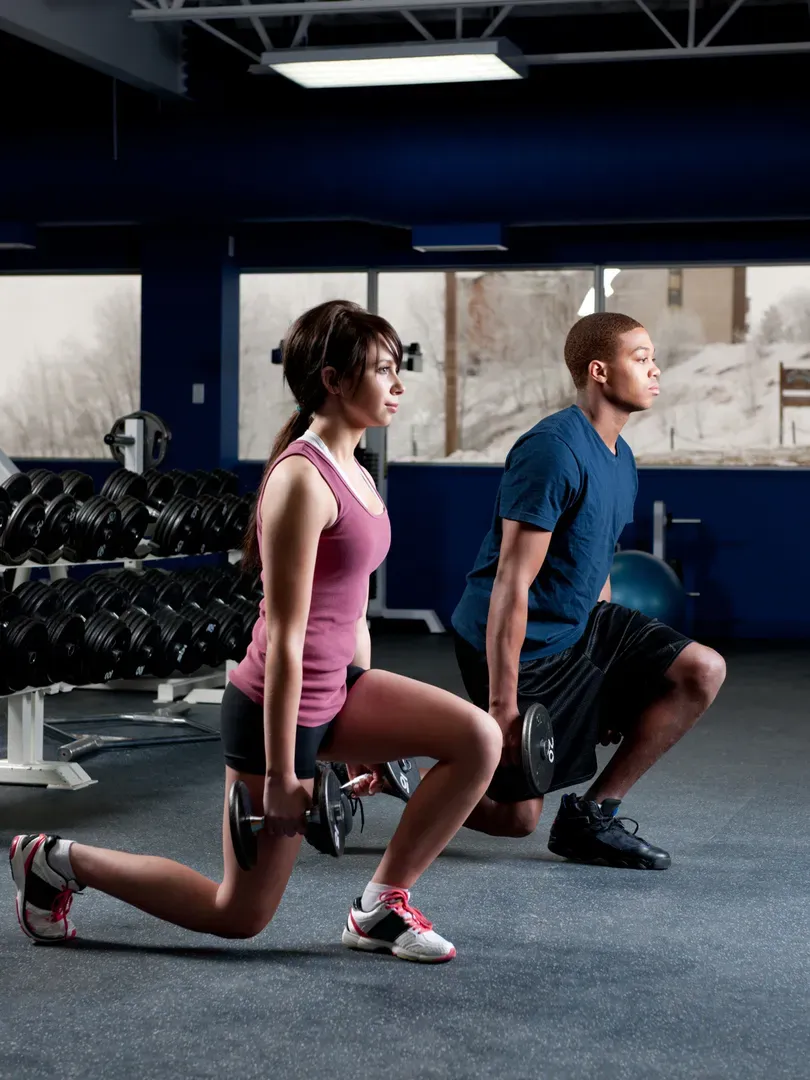Two people doing dumbbell lunges in a rec centre weight room
