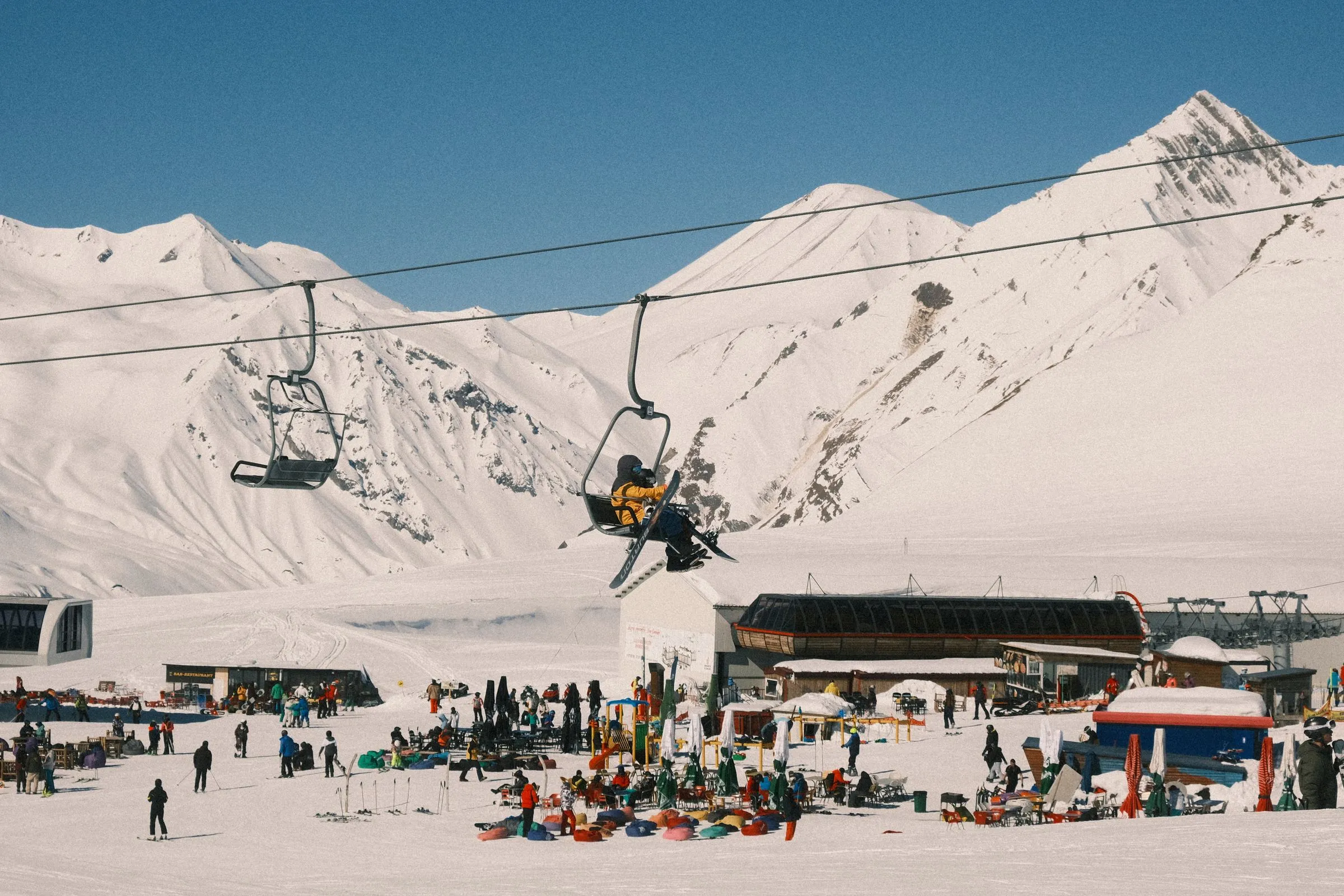 Skiers and snowboarders at a ski resort base area, with a chairlift carrying guests up the mountain